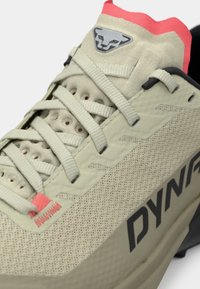 Αθλητικό παπούτσι σε ανοιχτό μπεζ χρώμα με υφασμάτινο πάνω μέρος με υφή, μαύρες λεπτομέρειες, ανοιχτό ροζ ταμπά πίσω και έντονο μαύρο λογότυπο "DYNAFIT".