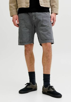 Man draagt grijze casual shorts met grote voorzakken, zwarte sokken en zwarte sneakers, staand tegen een eenvoudige lichte achtergrond.