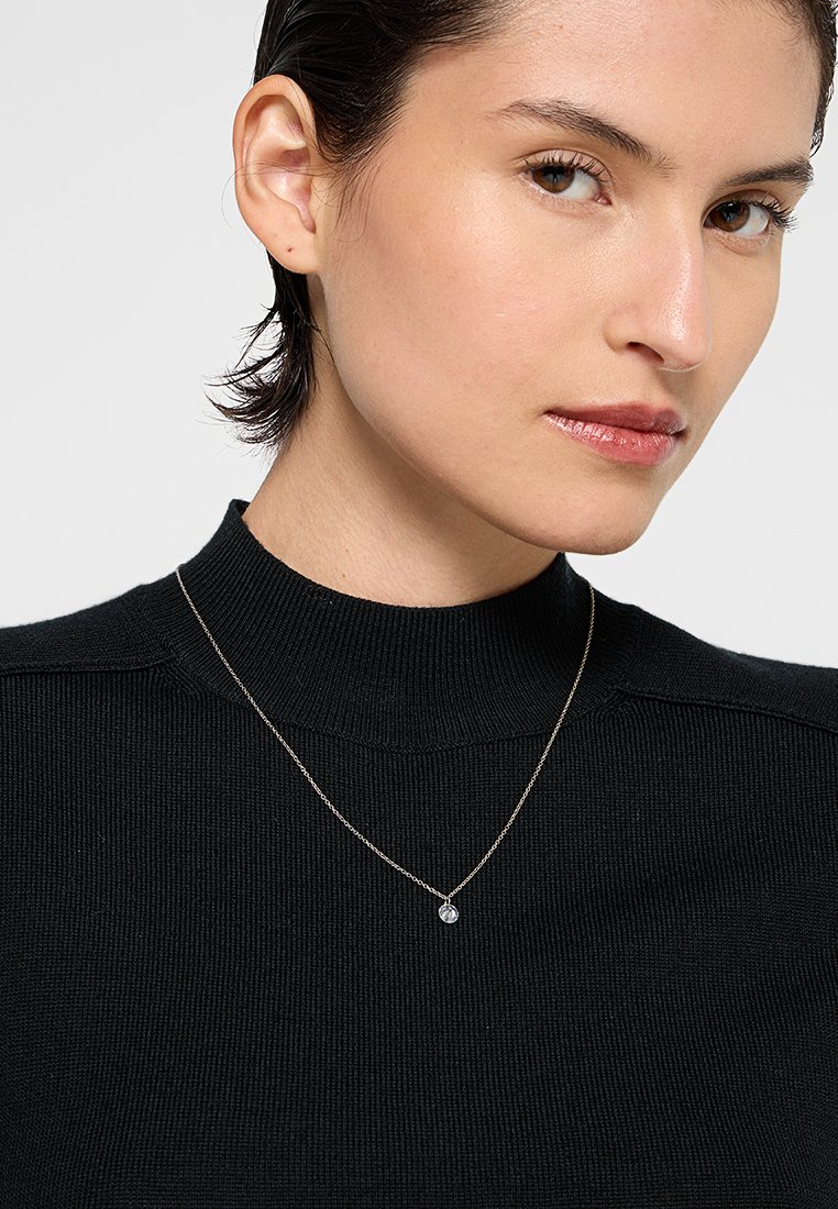 Collar de cadena de plata con un pequeño colgante redondo, llevado sobre un suéter de cuello alto de canalé negro. El tono de piel es claro y el fondo es liso.