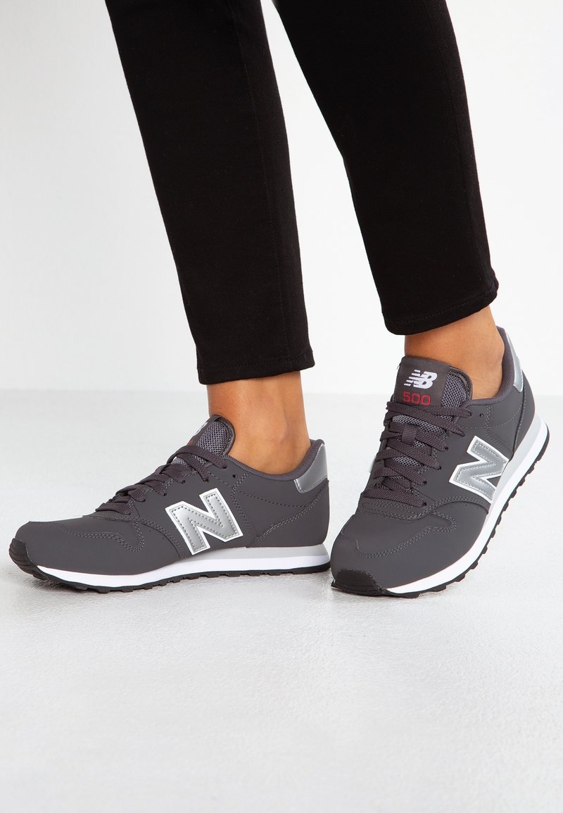 Pieds chaussés de baskets New Balance 500 grises avec semelles blanches et pantalon noir sur un fond blanc uni.