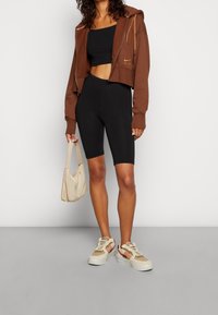 Brązowy skrócony hoodie z złotym zamkiem, czarny crop top, czarne szorty rowerowe, beżowa torebka i dwukolorowe sneakersy z różowymi akcentami.