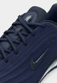 Ciemnoniebieskie buty biegowe z teksturowaną siateczką, płaskimi sznurowadłami i dużym błyszczącym logo w kształcie łyżwy po boku. Akcentowane detale w szwach.