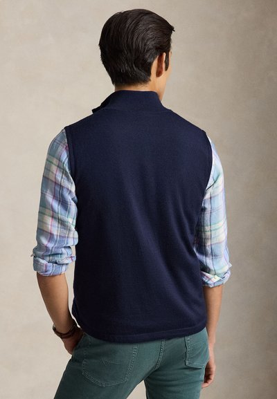 Vīrietis ar tumšiem matiem stāv ar seju pretēji, valkājot tumši zilu vestīti uz roll-up pasteļkrāsu pledzītas krekls un tirkīzzilām biksēm uz neitrālas fona.