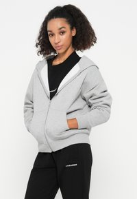 Grauer Reißverschluss-Hoodie mit gefüttertem Innenfutter, großer Kapuze und Fronttaschen. Getragen über einem schwarzen T-Shirt, kombiniert mit schwarzen Hosen mit weißem Branding.