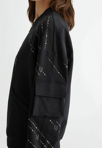 Felpa nera con chiusura a zip, tessuto testurizzato e borchie decorative argentate disposte in linee diagonali sulle maniche. Presente una grande tasca laterale.