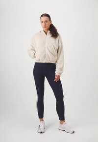 Kremna jopa z zadrgo iz flisa s sprednjim žepom, združena s temnimi legicami z belimi bočnimi progami in belimi športnimi copati.