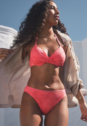 Vrouw in koraal gekleurde, gestructureerde bikini en gestreept overhemd staat buiten tegen een heldere blauwe lucht.