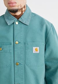 Veste boutonnée teal en coton, dotée de deux poches avant, d'un col à côtes et d'un patch logo Carhartt. Des boutons dorés ornent le design.