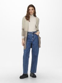 Béžový a hnedý pletený cardigan s otvoreným predným dielom, skombinovaný so svetlým rolákom a modrými džínsami s vysokým p�ásom, doplnený čiernymi topánkami.