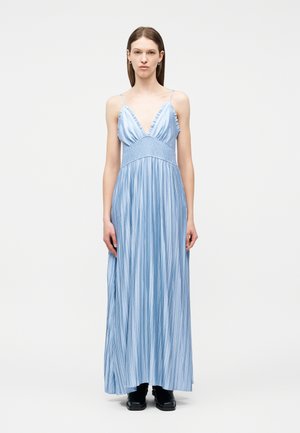 Femme debout portant une robe maxi plissée bleu clair sans manches avec de fines bretelles et des bottines noires sur un fond uni.