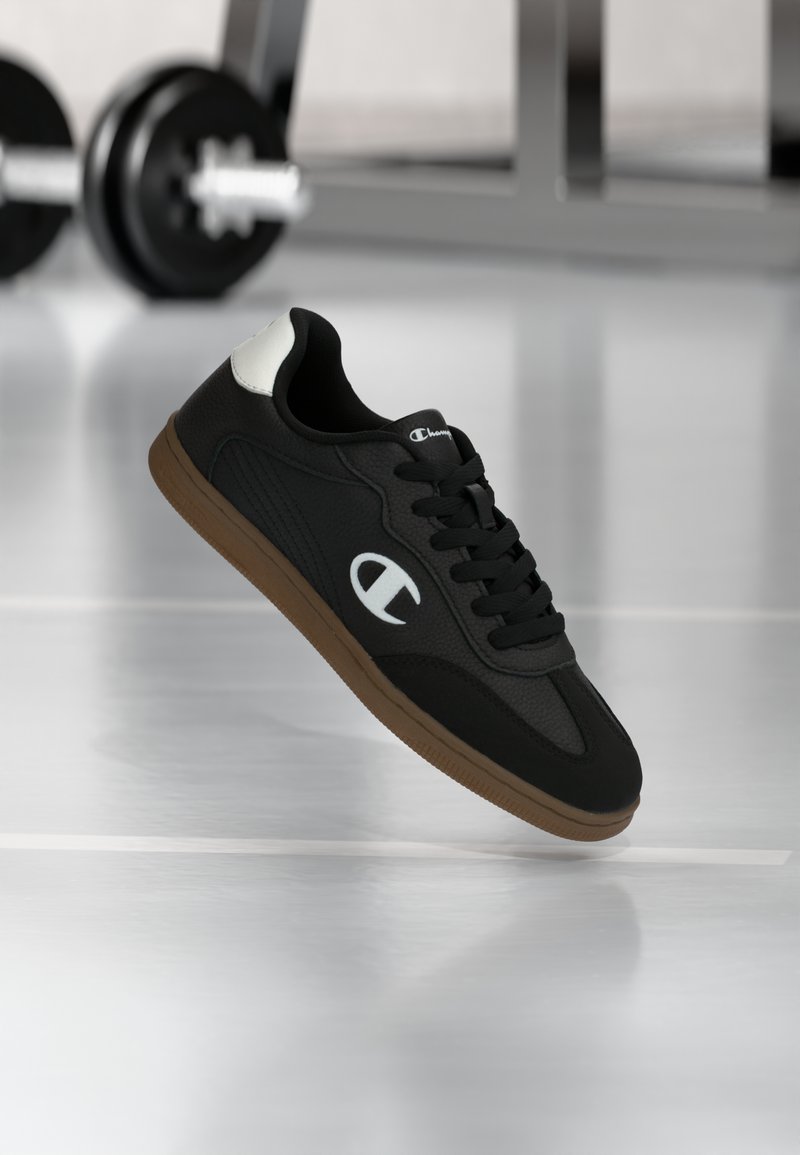 Svart sneaker med brun såle og hvit logo som svever over gymsgulvet med uskarpt vektløftingsutstyr i bakgrunnen.