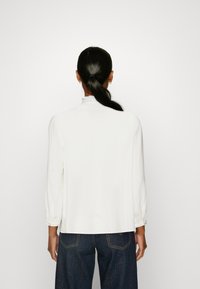 Blusa bianca a maniche lunghe con collo alto e polsini arricciati, abbinata a jeans in denim scuro. Tessuto morbido e vestibilità rilassata.