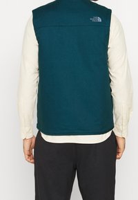 Tealfärgad väst i mjukt tyg, med hög krage, ärmhål och logotypdetalj på ryggen. Bärs över en ljusfärgad skjorta.