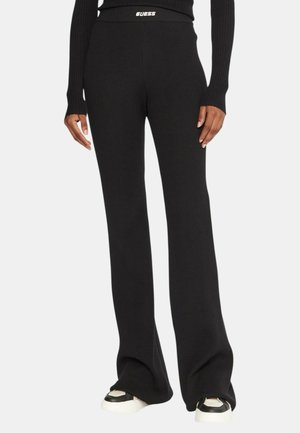 Persona che indossa pantaloni neri a coste a gamba larga e top a maniche lunghe coordinato, con scarpe da ginnastica bianche e nere, in piedi davanti a uno sfondo semplice.