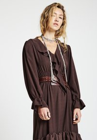 Braunes Kleid mit volants am Kragen und an den Ärmeln, tailliert und mit einem Gürtel mit silbernen Nieten versehen. Strukturierter Stoff mit ausgestelltem Saum.