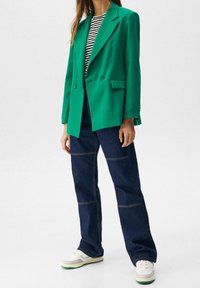 Blazer verde a medida con solapa, con dos botones; combinado con una camisa a rayas blanca y negra y pantalones de mezclilla de pierna ancha de color azul oscuro.
