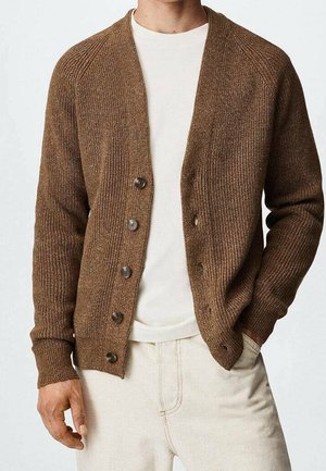 Mand iført brun ribstrikket cardigan med knapper over hvid skjorte og off-white bukser, med den ene hånd i lommen og den anden afslappet.