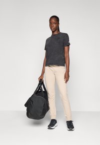 T-shirt noire à manches courtes avec une finition texturée, pantalon ajusté beige et chaussures de sport noires, tenant un sac de sport noir. Design simple.