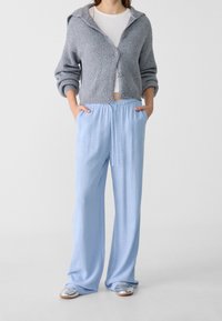 Gilet en maille gris avec capuche, fermeture à boutons et design court, associé à un pantalon large bleu clair et des chaussures argentées à enfiler.