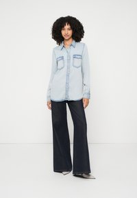 Light blue denim shirt with two front pockets, button-down front, and collar, paired with dark wide-leg jeans and gray pointed-toe shoes.