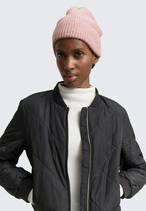 Woman wearing a pink knit beanie, black quilted jacket unzipped over a white ribbed turtleneck sweater, hands in jacket pockets.