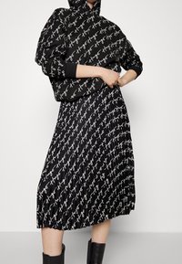 Černá mikina s bílým nápisem loga, spárovaná s plisovanou černou sukní se stejným vzorem loga. Hladká textura, ležérní design.