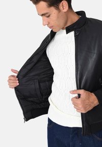 Giubbotto di pelle nera con fodera trapuntata, chiusura con zip e colletto alto. Presenta un maglione bianco a maglia cable sotto.