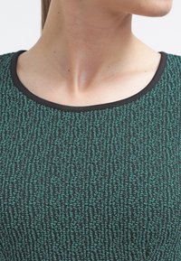 Vihreä ja musta kuvioitu toppi pyöreällä kaula-aukolla. Kangas sisältää tekstuuripainatuksen, jota korostaa musta vaatetus kaula-aukolla.