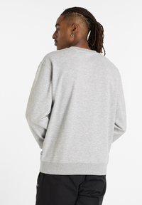 Man med flätat hår som är samlat bakåt, klädd i en ljusgrå rundhalsad tröja och svarta byxor, står vänd åt vänster med händerna i fickorna.