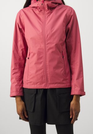 Vrouw draagt een roze regenjas met ritssluiting en capuchon, met ritssluitingen aan de zijzakken, gecombineerd met zwarte cargoshorts en zwarte leggings.