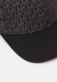 Gorra negra con una parte superior estampada que presenta la letra "K" en gris y un visera negra sólida. La tela parece suave con un diseño estructurado.