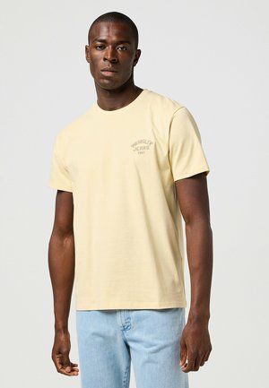 Camiseta de algodón amarillo claro con mangas cortas, cuello redondo y pequeño logo en el pecho. Llevada con jeans de mezclilla azul claro. Textura suave.