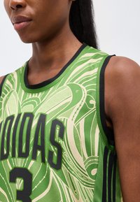 Prim-plan cu o persoană care poartă un tricou sport verde Adidas fără mâneci, cu margini negre și litere negre îndrăznețe.