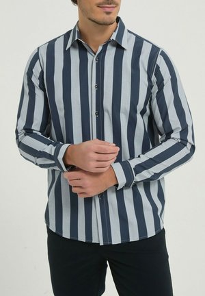 Homme ajustant le poignet d'une chemise à manches longues à rayures verticales larges bleu marine et gris clair, portant un pantalon noir.