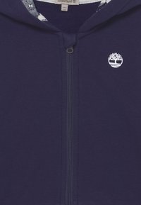 Felpa blu navy con zip frontale, logo ricamato bianco e fodera interna con motivo. Il tessuto sembra morbido e texturizzato.