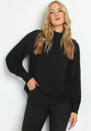Zwarte lange mouwen top met hoge kraag, gestructureerde stof en licht gepofte mouwen. Gekocht met zwarte jeans, voorzien van zijzakken.