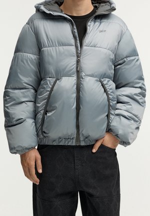 Homme portant une veste matelassée gris clair avec fermeture éclair et poches à l'avant, et un pantalon noir à motifs, une main dans la poche.