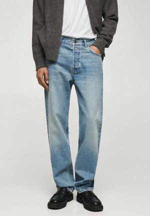 Homme portant un jean bleu clair à coupe droite, un t-shirt blanc, un pull gris à fermeture éclair, et des chaussures noires à semelle épaisse, debout avec une main dans la poche.