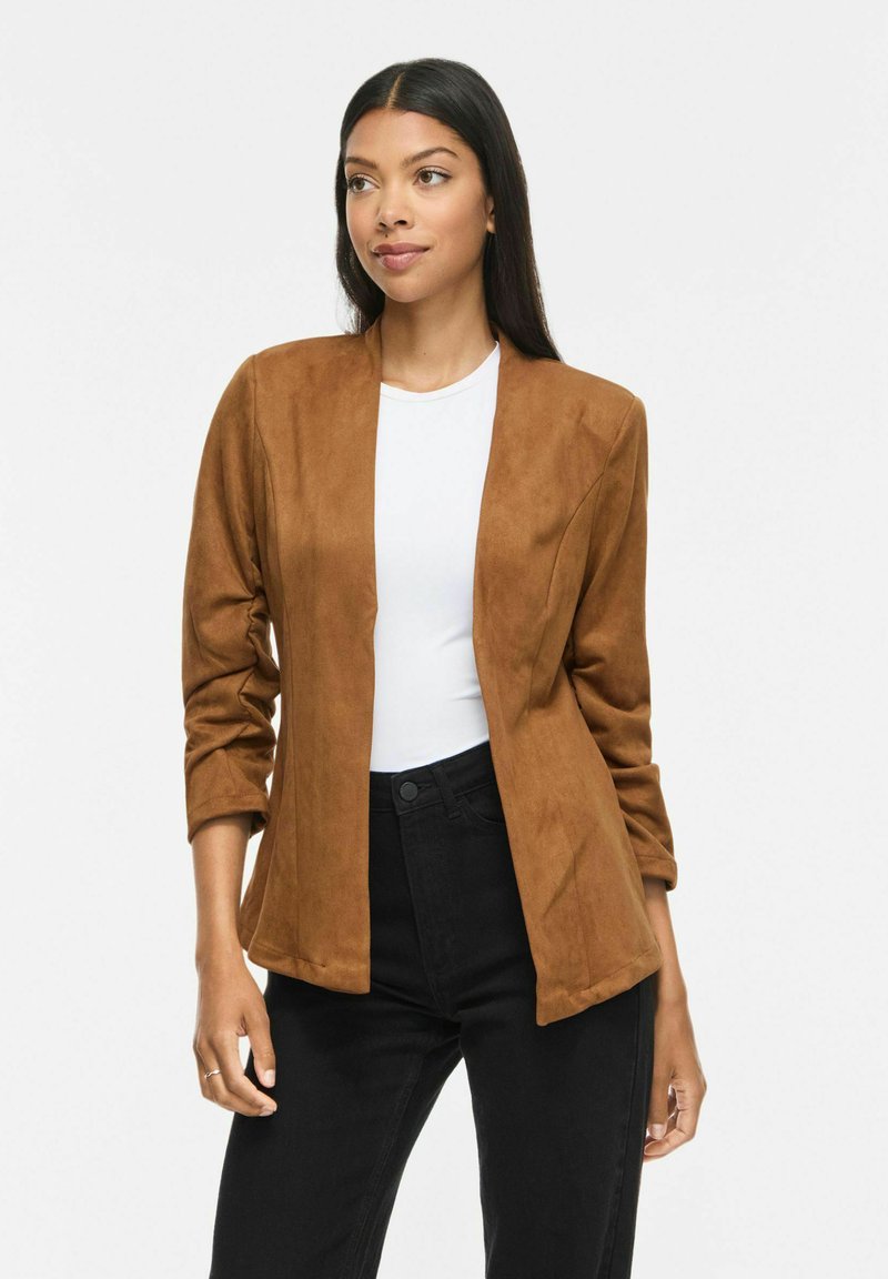 Femme aux longs cheveux foncés portant un blazer marron ouvert, une chemise blanche et un pantalon noir, debout devant un fond blanc uni.