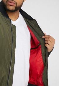 Giubbotto bomber verde oliva con un'esterno liscio, chiusura a zip e fodera in raso rosso. Presenta un colletto e polsini a coste.