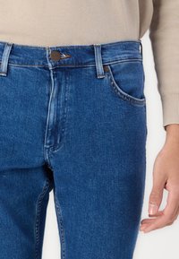 Nærbilde av en person som har på seg blå denimjeans og beige genser, viser midje, lomme og høyre hånd ved siden.