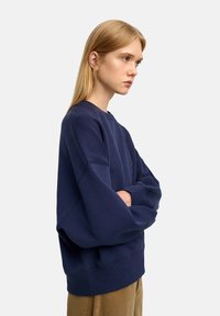 Granatowy sweter w luźnym kroju, z okrągłym dekoltem, obniżonymi ramionami i prążkowanymi mankietami. Wykonany z miękkiej bawełny, o minimalistycznym wzornictwie.