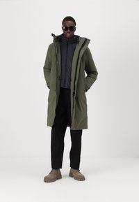 Olivgrüner Parka mit Kordelzugkapuze, über einer schwarzen Zip-Jacke getragen. Kombiniert mit schwarzen Hosen und sandfarbenen Schnürboots.