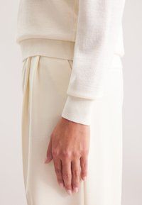 Maglione di maglia crema con polsini a coste, abbinato a pantaloni plissettati color crema chiaro. Mano appoggiata sulla coscia, texture della pelle liscia visibile.