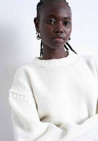 Femme à la peau foncée et aux cheveux tressés portant un pull en maille blanc et des boucles d'oreilles créoles argentées avec des accents verts, sur un fond clair uni.