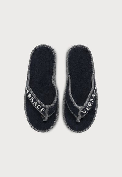 Paire de sandales tongs en tissu éponge noir avec garniture blanche et texte « VERSACE » sur les brides en tissu sur fond blanc uni.