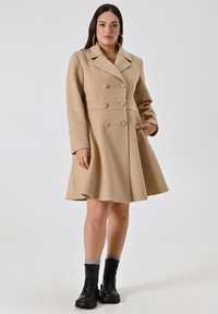 Cappotto beige doppiopetto con gonna svasata, realizzato in un materiale morbido. Presenta grandi bottoni, colletto a revers e tasche laterali. Stivaletti neri con lacci.