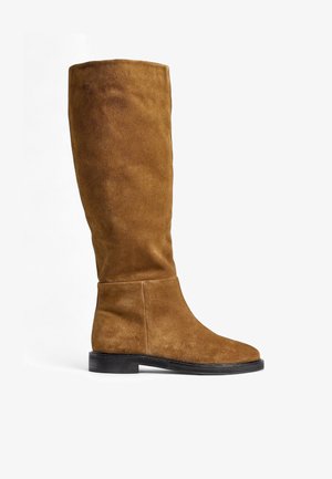 Bottes en suede marron montantes avec un bout rond, une semelle plate et un design sans couture, présentant une finition cousue le long de la cheville et du haut.