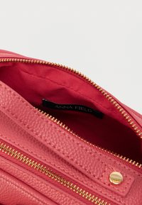 Ροζ τσάντα από δερματίνη με υφή, με χρυσά φερμουάρ και μεταλλικά στοιχεία. Εσωτερικά επενδεδυμένη με κόκκινο ύφασμα, με ετικέτα μάρκας ορατή μέσα.