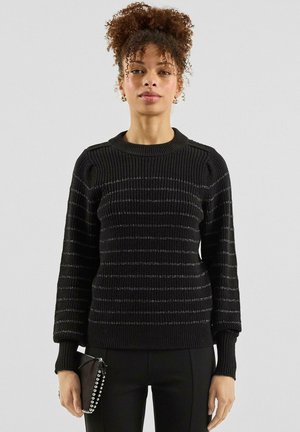 Jeune femme aux cheveux bouclés portant un pull noir côtelé avec des rayures argentées, un pantalon noir, et tenant une petite pochette noire cloutée.